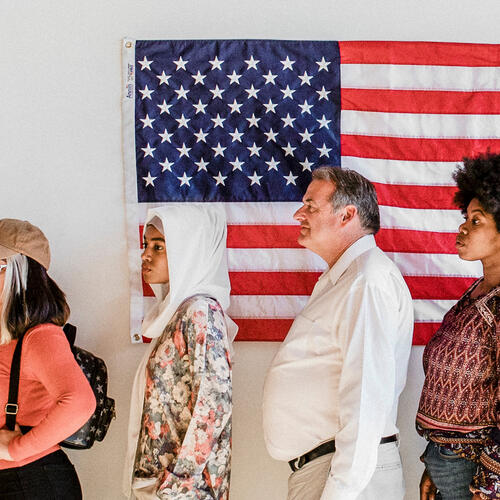 Photo: Diverse group of people standing in line with an American flag displayed behind them