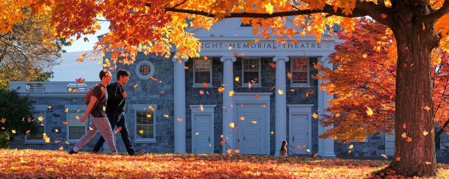Photo of two students walking in front of a stone building with pillars and a tree