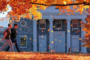 Photo of two students walking in front of a stone building with pillars and a tree